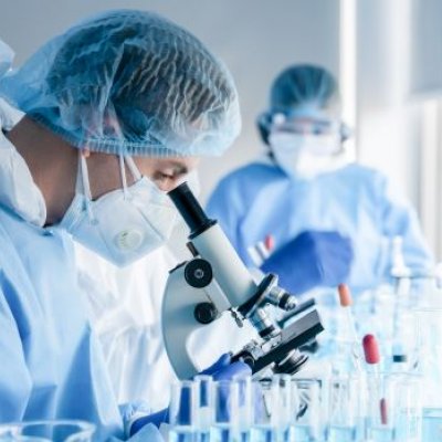 A scientist in cap, gown and mask stands in profile looking down through a microscope as a second scientist looks on in the background. They are in a lab.